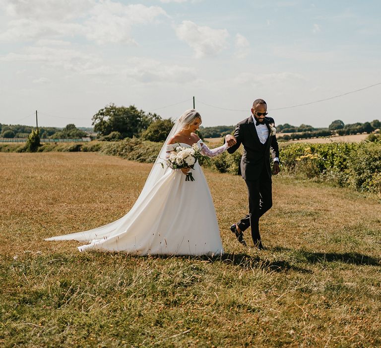 Bride in princess wedding dress and detachable lace sleeves walks alongside her groom in black-tie across golden fields 