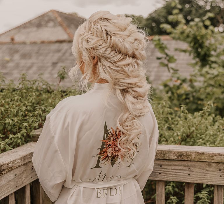 Bride in a personalised white robe with her platinum blonde hair in a pulled apart boho fishtail braided hair in an intricate style looking out at the view 