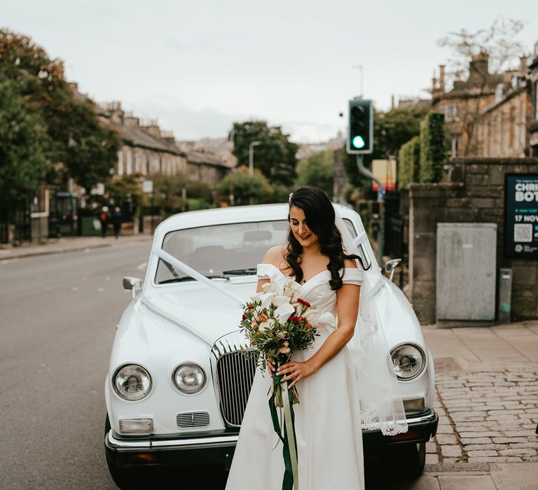 Bride carries orchid bridal bouquet and leaves white vintage wedding car with ribbon to the front 