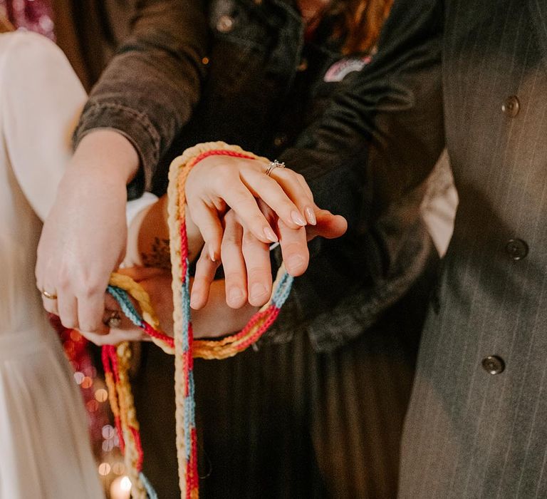 Celebrant in black velvet outfit doing handfasting ceremony with multicoloured knitted tie before doing jumping the broom ceremony at glam grunge Sheffield wedding 