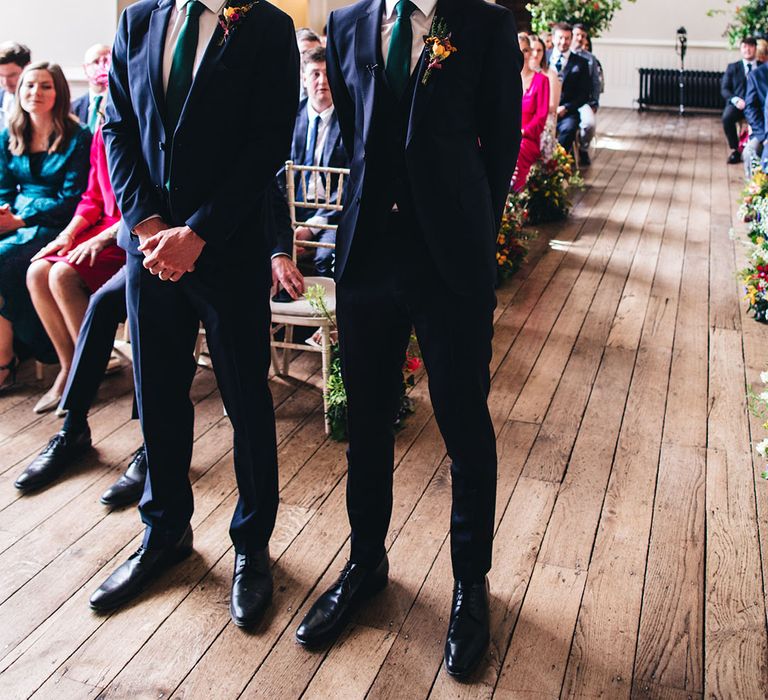 Groom stands in a three piece navy suit and green tie with colourful buttonhole at the altar with the best man also in a navy suit and green tie 