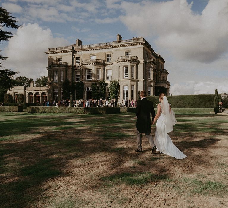 Bride in floor-length cathedral veil walks alongside her groom in Morning suit toward Hedsor House 