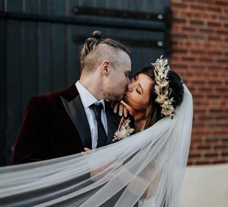 Groom in burgundy velvet suit jacket kissing the bride who wears a veil and boho dried flower crown 