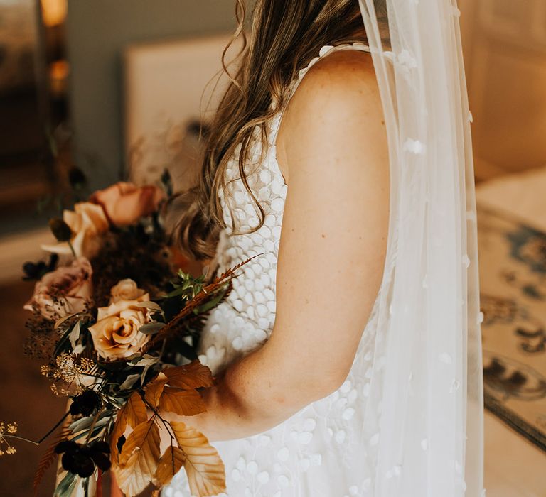 Bride in a Made With Love wedding dress and matching veil with he hair in a braided half up half down hairstyle holding a neutral palette bouquet for an autumn wedding 