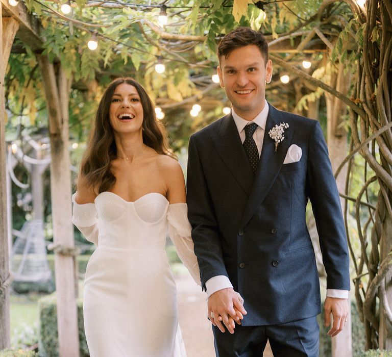 Groom in double breasted navy suit with spotted tie walking hand in hand with the bride in Jimmy Choo pearl shoes and modern wedding dress