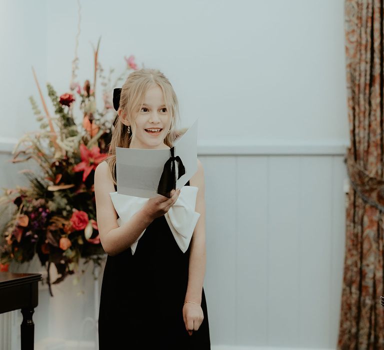 The bride and groom's daughter in a black dress with large white bow and tights with metallic shoes reads out a wedding reading for the ceremony