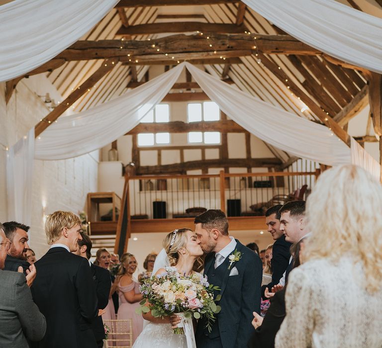 White drapery and fairy lights decorate the Dove Barn wedding venue with the bride and groom sharing a kiss as they walk back down the aisle 