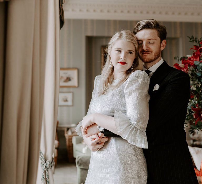 Groom in pinstriped suit embracing the bride from behind wearing metallic wedding dress with shoulder pads and ruffle sleeves 