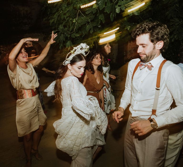 Bride in lace wedding dress, pearl earrings and bridal crown dances at wedding reception in Italy with groom in burnt orange braces and hwite wedding shirt