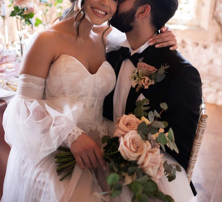 Bride in off shoulder wedding dress with detachable puff tulle sleeves and embroidered rose 3d appliqué details holding blush rose and eucalyptus bridal bouquet embracing groom in classic black tuxedo with garden rose and eucalyptus boutonniere 