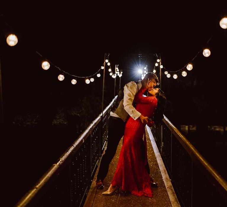 Groom in grey waistcoat dancing and kissing bride in red lace wedding dress with long sleeves on a bridge under wedding lanterns at The Orangery Maidstone