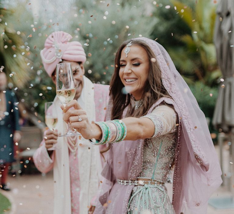 Bride holding up a glass of champagne during colourful confetti exit at three day multicultural destination wedding 