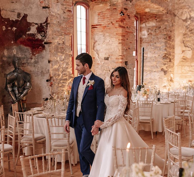 Groom in blue suit with a red tie walking though the wedding venue with the bride in an Eva Lendel wedding dress 