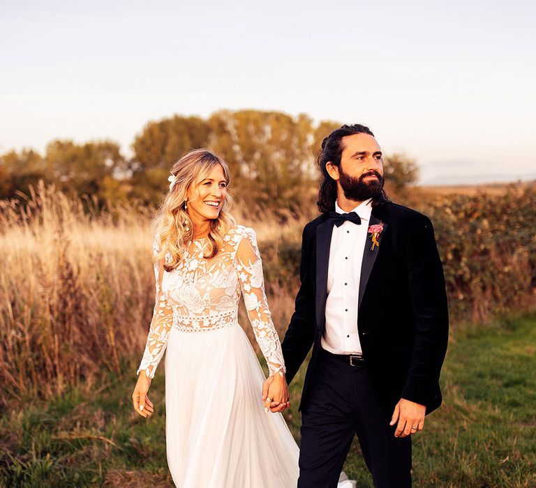 Groom in black tuxedo walking hand in hand with bride in long sleeve wedding dress 