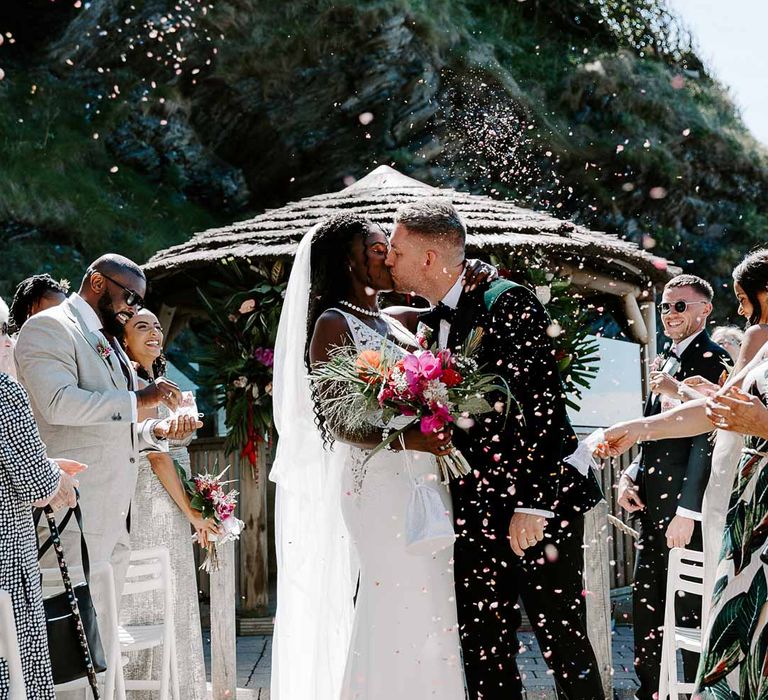 Groom in bottle green velvet grooms blazer, colourful tropical boutonniere and black suit trousers kissing bride in v neck sleeveless lace Pronovias wedding dress and cathedral length veil doing colourful confetti exit at Tunnels Beaches 