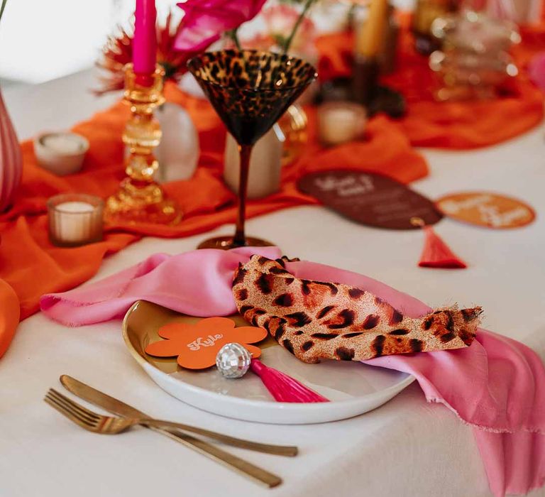 Leopard print wedding fabric fabric on top of pink wedding napkin on retro wedding tablescape with pink tapered candles, orange table runner, mismatched single flower vases, colourful flower centrepieces and large colourful flower arrangements 