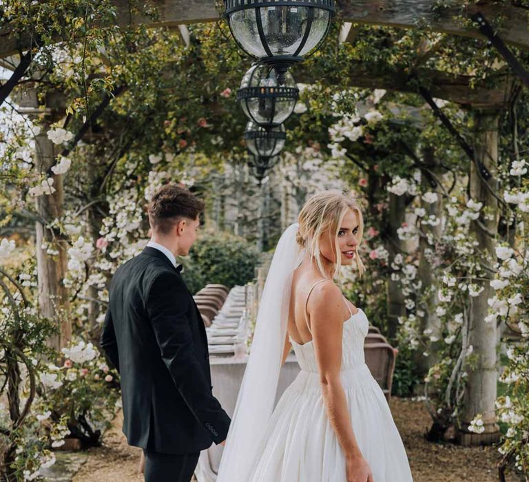 Groom in classic black tie tuxedo walking with bride in Watters strappy sleeveless princess wedding dress and chapel length wedding veil by outdoor wedding tablescape at Euridge Manor wedding 