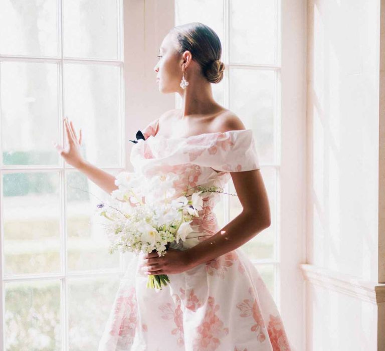 Bride wearing off the shoulder white and pink floral patterned bridal gown with black bow and front slit holding bridal bouquet with white poppies, light pink Persian buttercups, baby's-breath and foliage looking out of the window at Settrington Orangery wedding venue