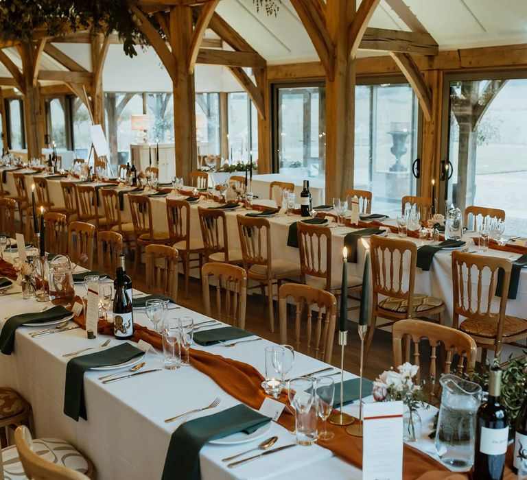 Winter themed classic wedding tablescape with white tablecloth, burnt orange wedding tablerunner, forest green wedding napkins, forest green tapered candles and bottles of wine with suspended foliage and fairy lights decor at wedding