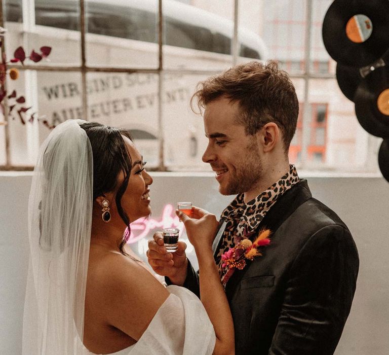 Groom in wet look black grooms suit with leopard print shirt and pink and orange boutonniere standing by vinyl wedding arch with bride in off shoulder tulle short wedding dress with church length veil doing wedding shots 