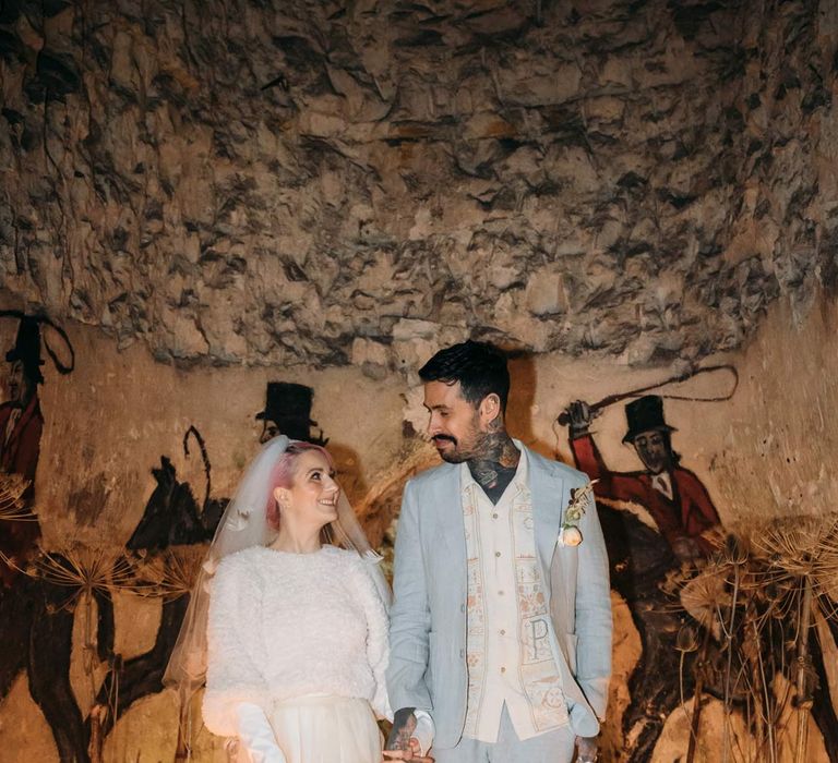 Groom in baby blue linen suit, personalised embroidered linen shirt and garden rose and pampas grass boutonniere with bride in sheer overlay wedding dress and fluffy bridal coverup surrounded by tapered candles in Margate Caves