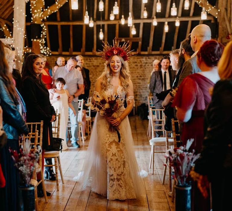 Bride in off shoulder lace wedding dress with corset style top and red rose and gold halo bridal crown holding red rose, pampas grass and dried flower bridal bouquet walking down the aisle at Southlands Barn