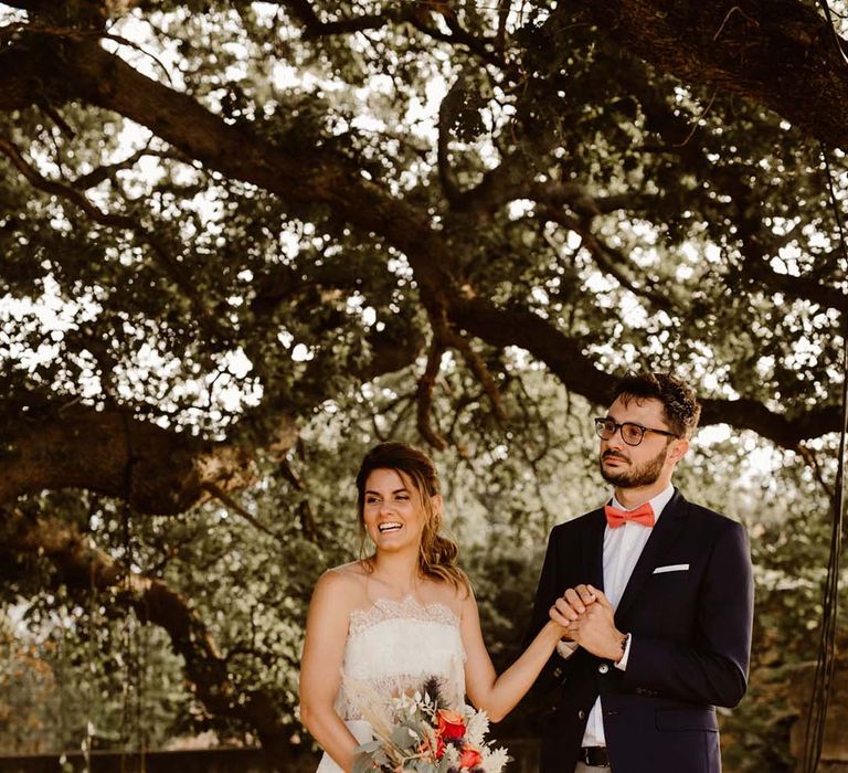 groom in grey chinos navy blazer and orange bow tie holding hands with his bride in a strapless lace wedding dress