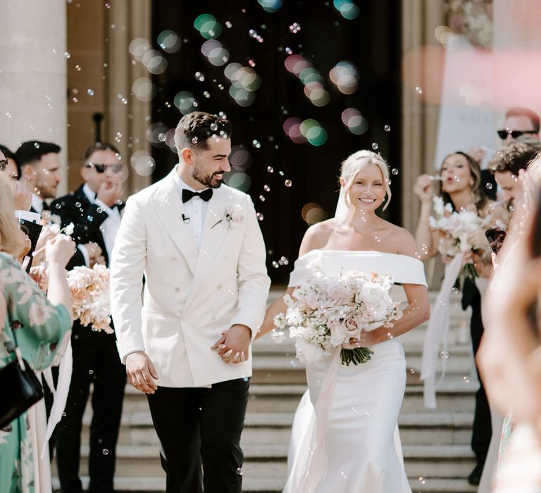 Bubble exit from the ceremony alternative to confetti with bride in off the shoulder wedding dress and groom in white suit jacket 