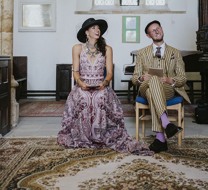 Bride in pink bohemian wedding dress sits next to groom in pinstripe suit at church wedding ceremony