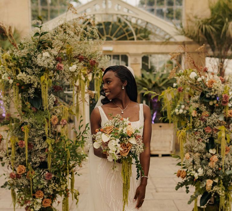 Bride stands in front of wedding flower columns with green hanging amaranthus 