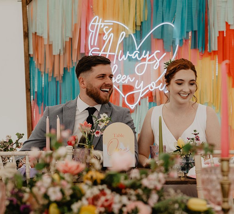 The bride and groom sit at their wedding table decorated with a pink neon sign and colourful streamers 