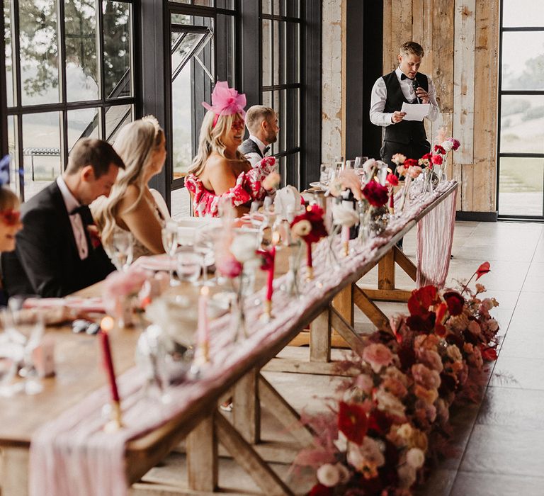 The bride, groom and wedding guests sit on long wooden table listening to the wedding speech 