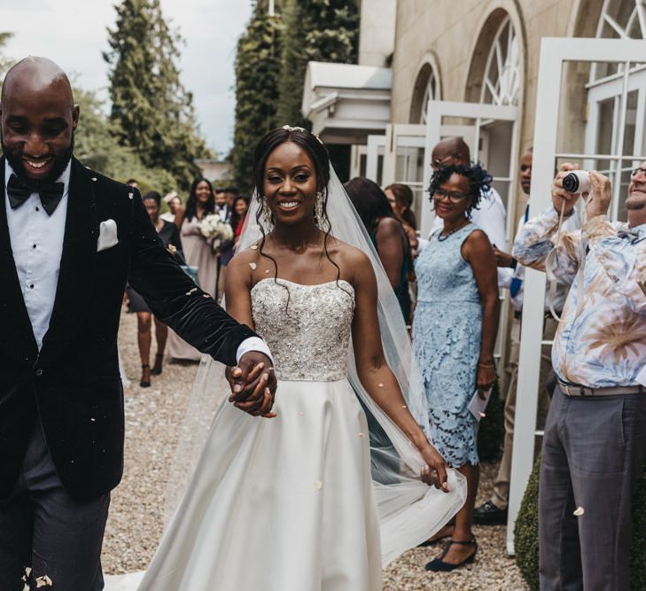 Black groom in a velvet tuxedo and bow tie holding his brides hand in a strapless Oleg Cassini wedding dress during the confetti moment 