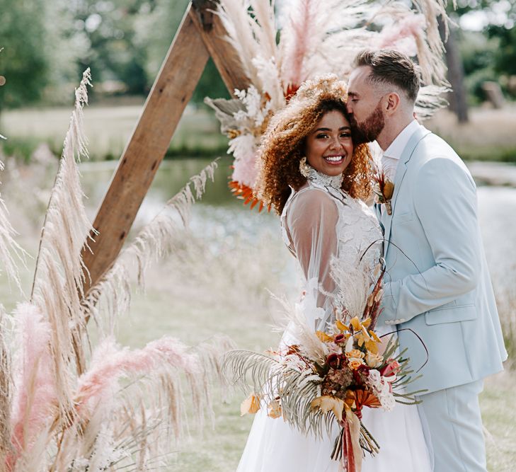 Groom kisses bride whilst she holds dried floral bouquet and hair blows in the wind