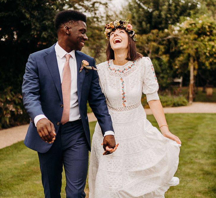 Groom in a navy suit with red patterned tie, smiling at his bride in a laser cut dress with colourful embroidery detail 