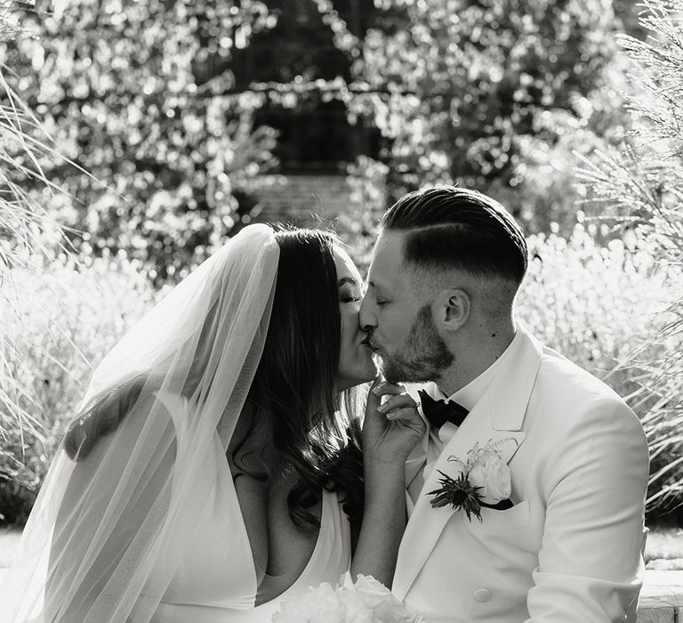 Bride in V neck Made With Love wedding dress and veil holding white rose bouquet sits outside with groom in white double breasted tuxedo jacket