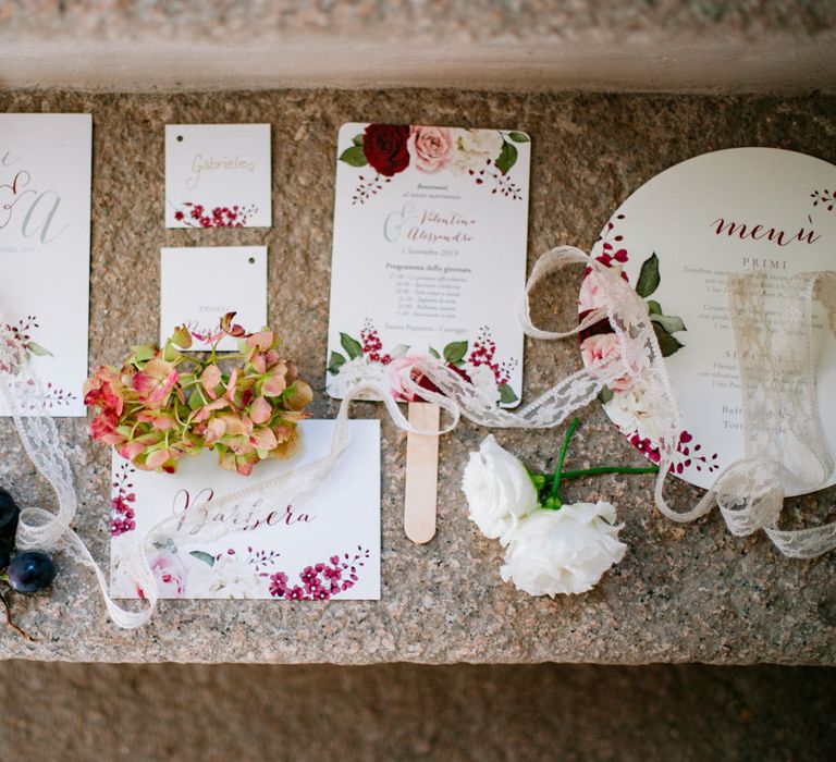 Red and white wedding stationery decorated with roses, next to lace ribbon and Italian grapes