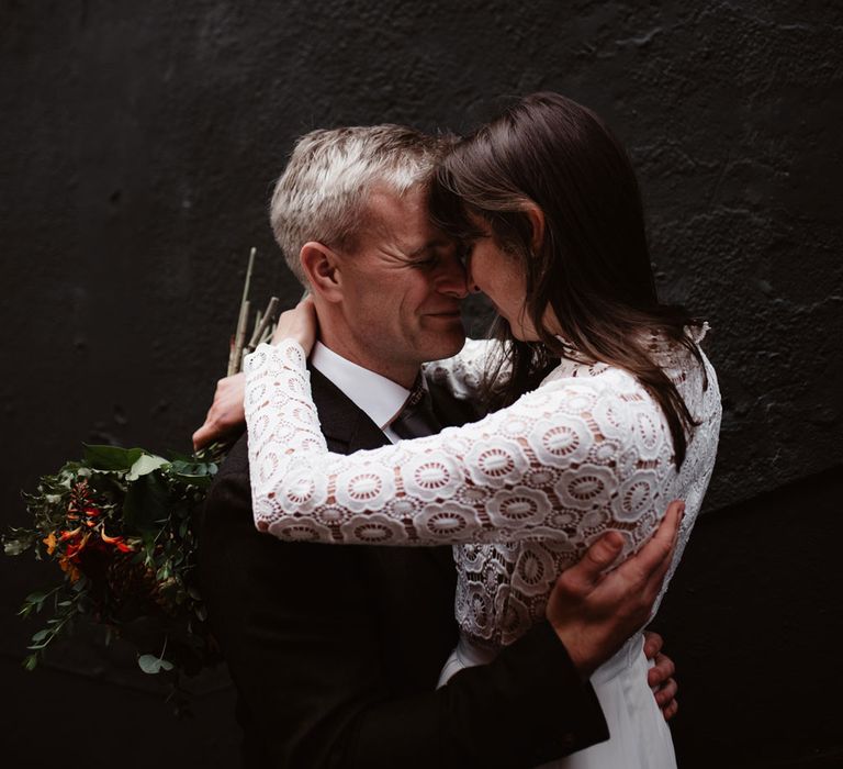 Bride in long sleeved lace top Self Portrait wedding dress hugs groom in brown woollen blazer and grey trousers on steps by black wall in Bristol