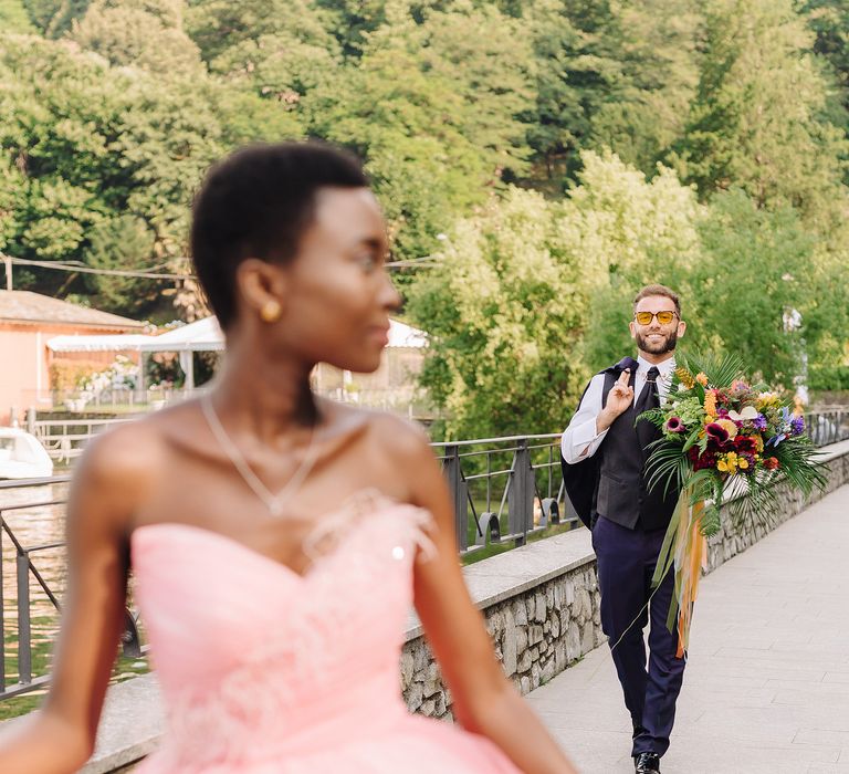 Bride wears pink bridal gown with sweetheart neckline as groom walks toward her holding bouquet