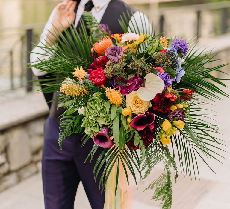 Groom holds colourful bouquet with green foliage and tied with iridescent ribbon