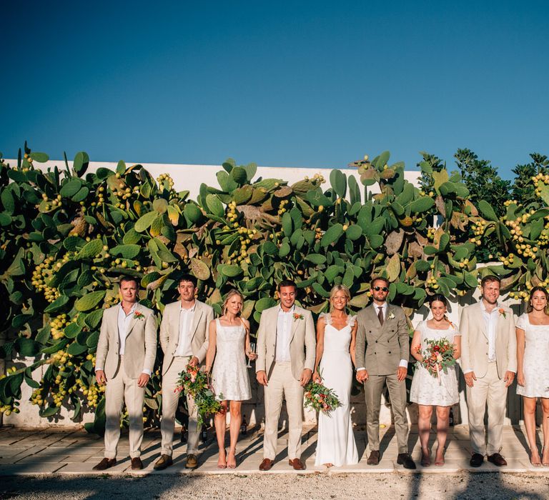 The bride, groom, bridesmaids and groomsmen standing together in the sun in front of a cactus wall