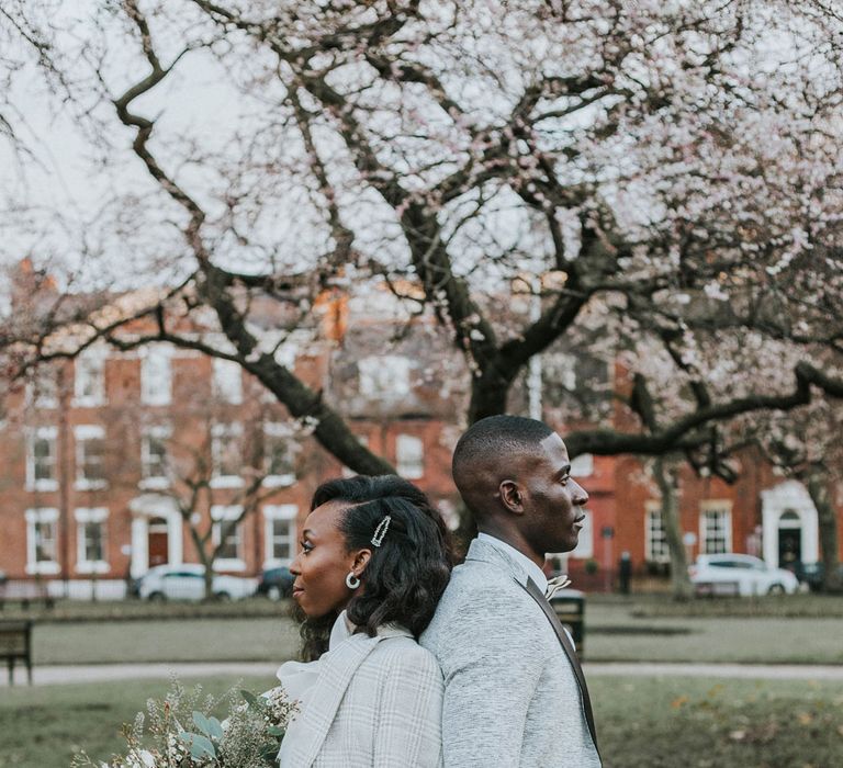 Bride in white satin dress and light grey checked blazer holding white and green wedding bouquet stands back to back with groom in grey jacket and black trousers in a park after Bridge Community Church wedding