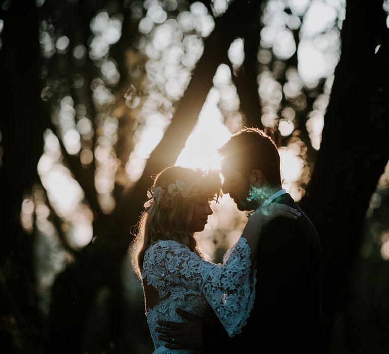 Bride in long wide sleeved lace wedding dress with curled hair and flower crown stands in forest with hands resting on groom in navy suit with bow tie at enchanted forest wedding