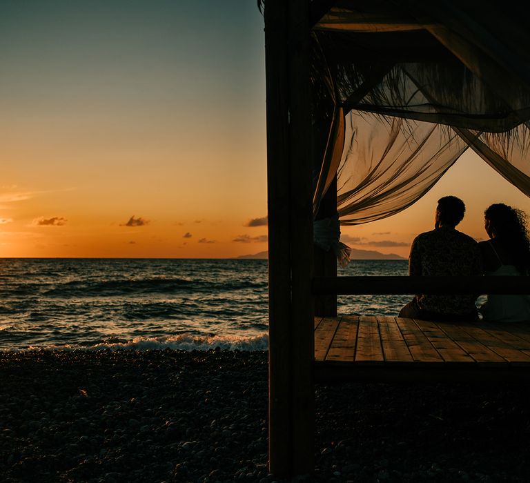 Bride & groom sit on day bed on the beach in Santorini watching the sun set