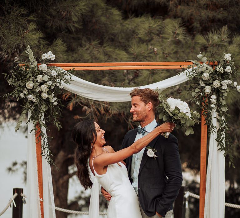 Bride and groom share a laugh under the wooden arch decorated in white drapes and flowers