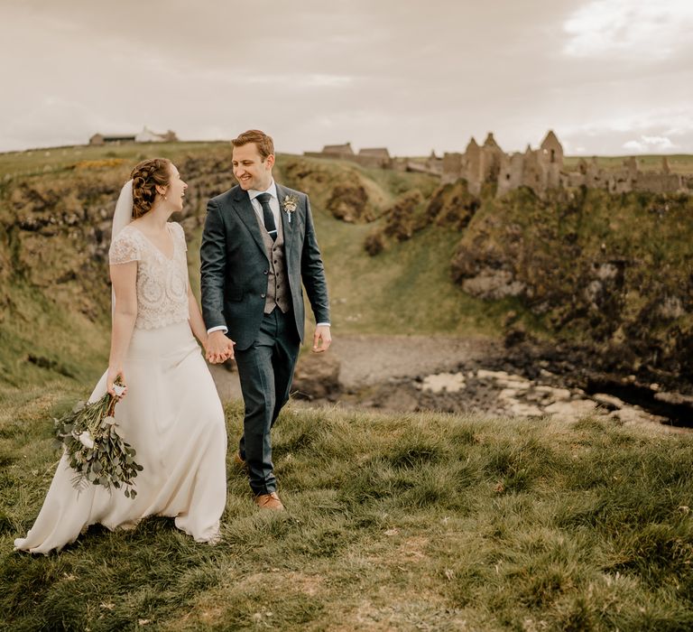 Bride in lace top capped sleeve wedding dress and satin skirt holds mixed bouquet whilst holding hands with room in navy suit walking along clifftop with Dunluce Castle in the background