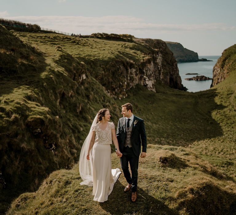 Bride in lace top wedding dress with satin skirt and veil holds hands with groom on clifftop after Dunluce Castle wedding