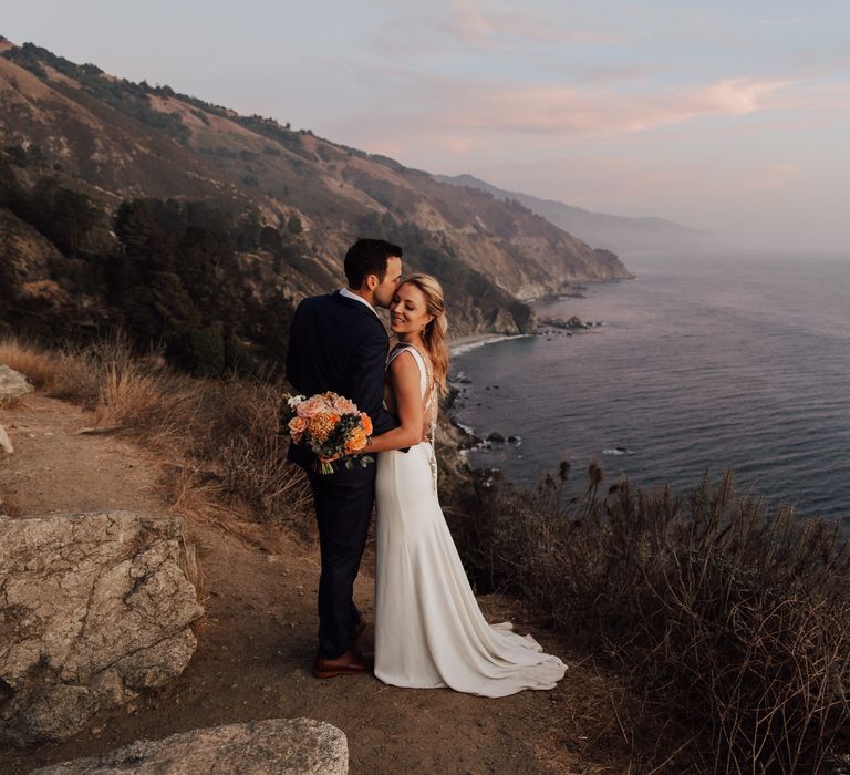 Bride & groom stand on the cliffside as the sunsets behind the ocean