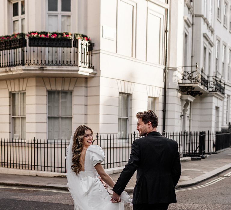 Bride with long wavy hair wearing a short wedding dress with open back holding hands with her husband in the streets of London 