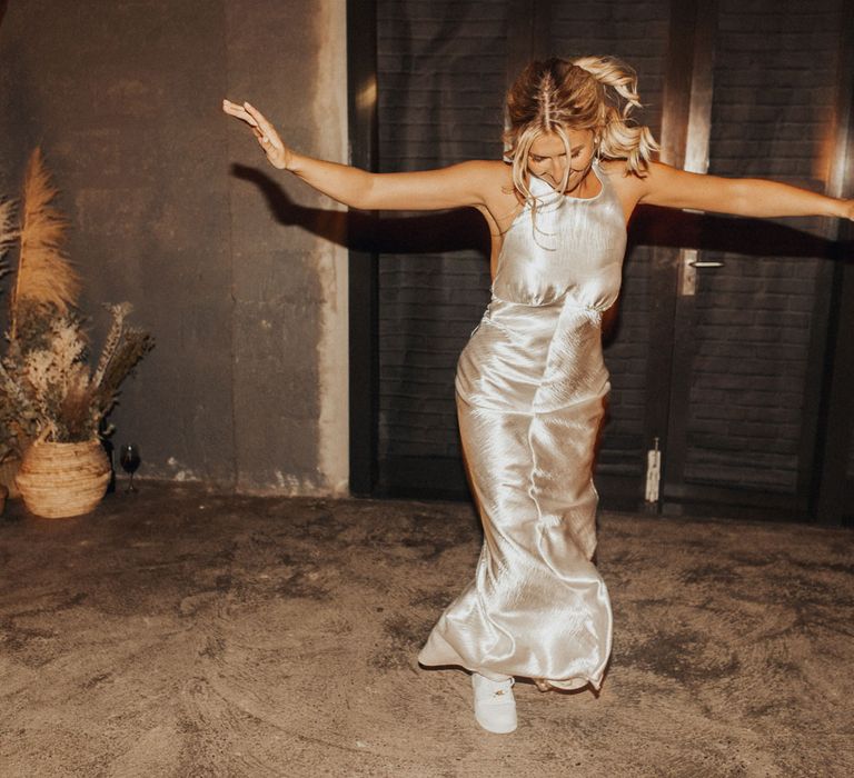 Bride with curled ponytail in silver satin dress and white air force trainers dances in the hut at Anran Devon for wedding evening party